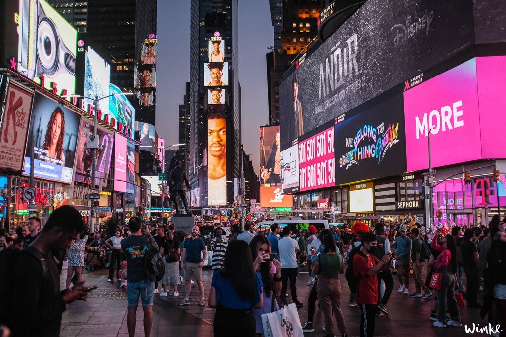 Overweldigend uitzicht op Times Square 's nachts, vol met felle billboards, druk verkeer, diverse mensen inclusief personen in kostuums en zwervers, een sfeer van muziek en bruisend stadsleven, terwijl een onbekend persoon op de trappen zit met een ongezoete ijsthee van McDonald's.