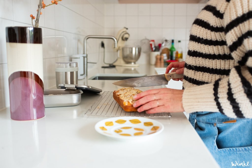 Een persoon in een gestreepte trui en blauwe spijkerbroek snijdt een zelfgebakken brood op het aanrecht van de keuken. Naast hen staan stijlvolle keukencontainers van Brabantia en een doorzichtige lunchbox met een sandwich, wat duidt op de voorbereiding van een maaltijd om mee te nemen.