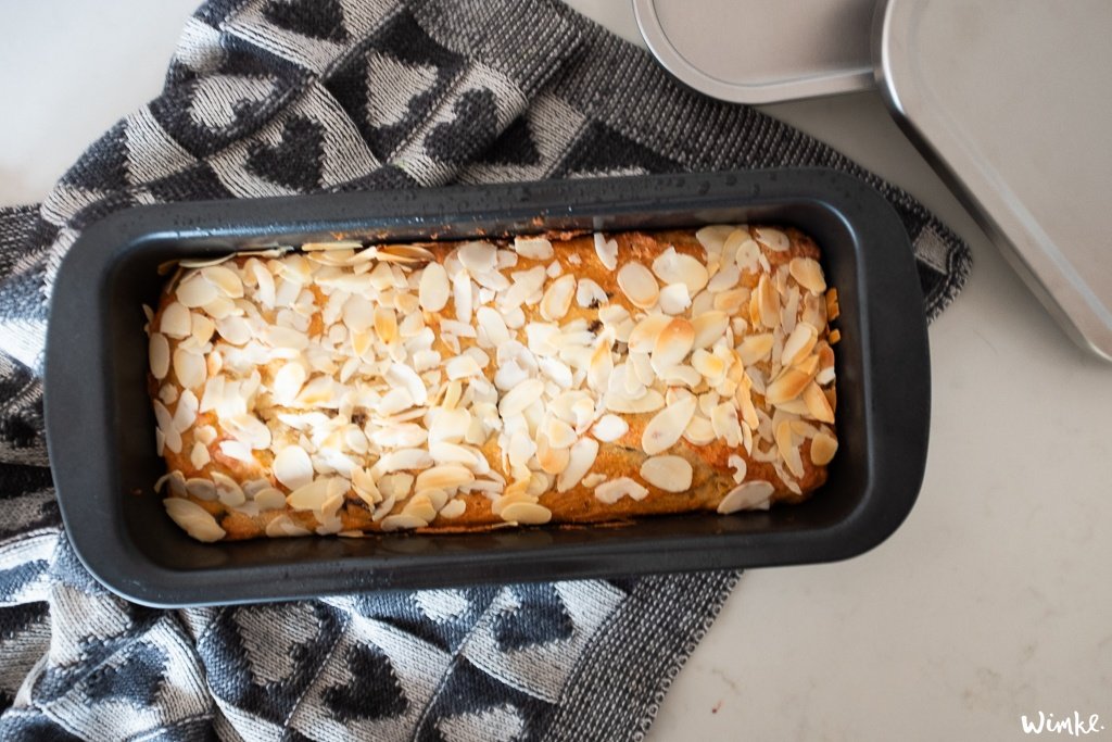 Een versgebakken brood met amandelschaafsel in een Brabantia broodvorm, rustend op een afkoelrek met een keukendoek eronder. Het brood is goed gerezen en goudbruin, wat wijst op een heerlijk zelfgemaakt recept dat klaar is om verpakt te worden voor een maaltijd.