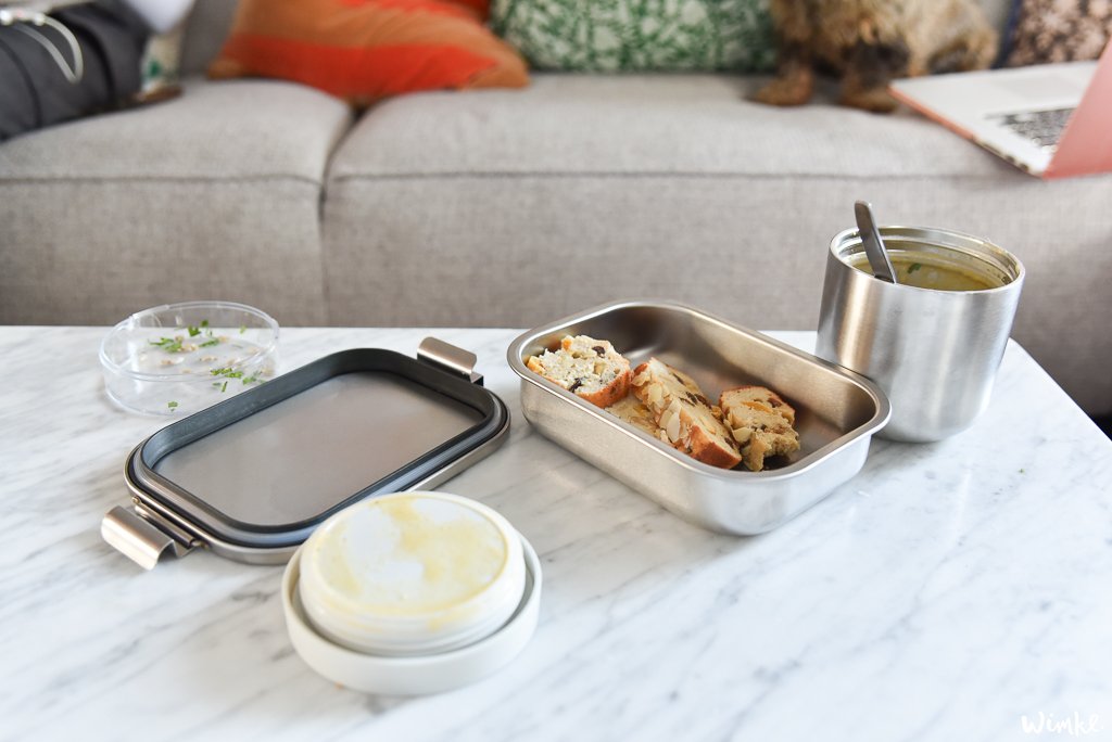 Een moderne thuiswerkopstelling met Brabantia's Make & Take op een marmeren aanrecht. Een sandwich in een roestvrijstalen lunchbox, een doorzichtig bakje met kruiden, een soepthermos met lepel en een laptop op de achtergrond suggereren een productieve omgeving voor thuiswerkers.